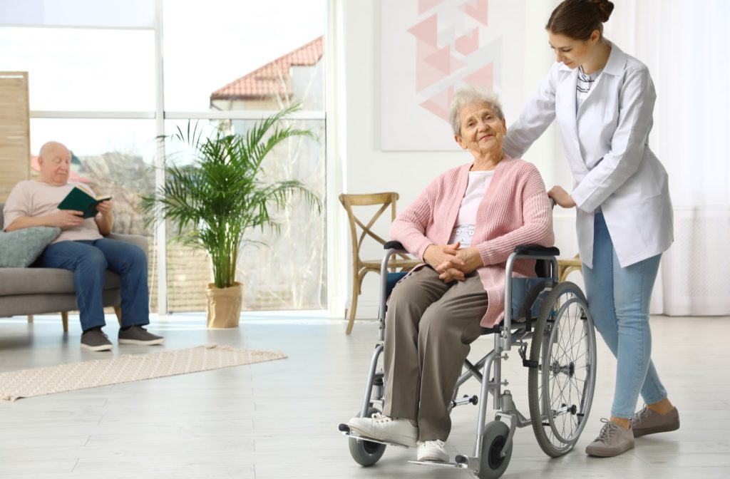 Nurse assisting elderly women in wheelchair at senior community with a man reading book on chair in background
