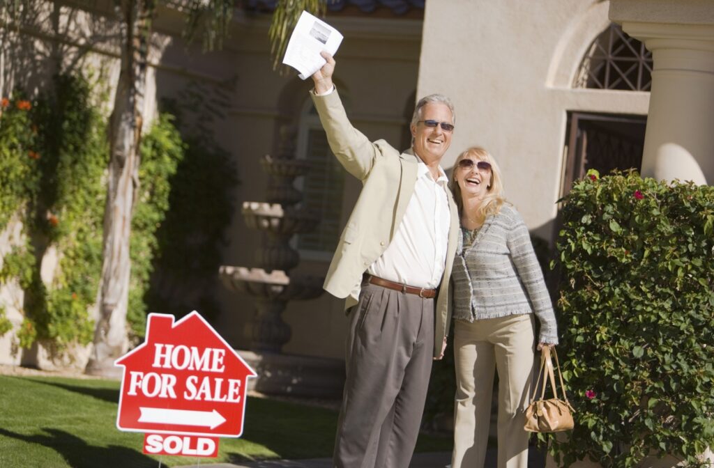 A happy senior couple stand outside their home, smiling with a SOLD sign in the lawn and the paperwork in their hands.