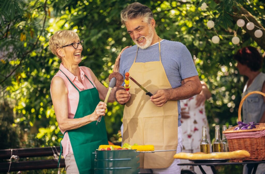 Two older adults laugh and joke together while preparing food as part of a public outdoor community celebration.