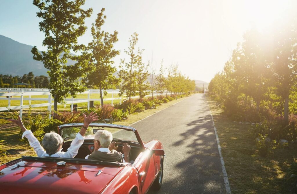 A senior couple in a red convertible drives down a paved countryside road into the sunset