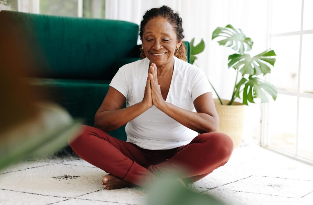 A senior sits cross-legged on the floor, holding their hands together to meditate in a simple yoga pose