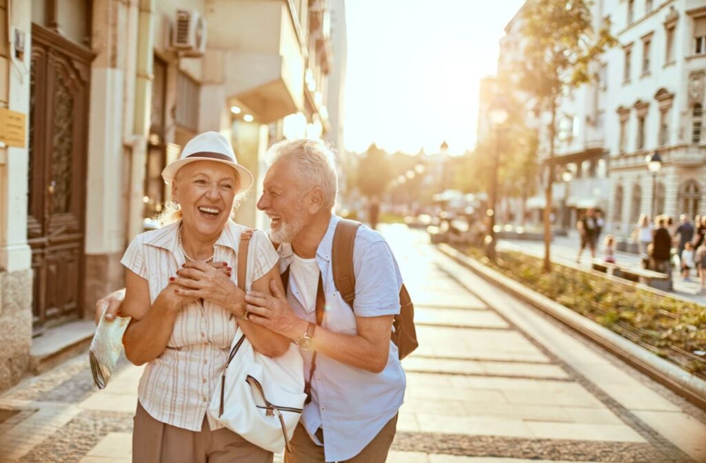 An older couple laughs while walking arm-in-arm as tourists down a warm sunlit street in a new city